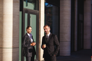 Fototapeta na wymiar Portrait of two confident business people in suits, young businesswoman and middle aged businessman, looking at camera and smiling while posing near office building with takeaway coffee cups