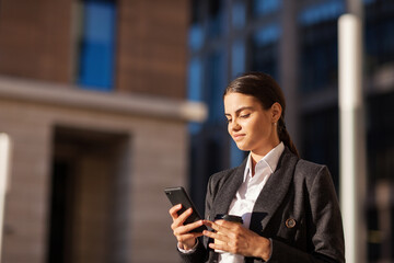 Side view of brunette young woman in suit text messaging on cell phone standing outdoors with takeaway coffee cup in her hand, copy space to left