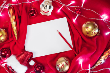 A clean white sheet of paper and a red pencil lie on the blanket, and next to them are red and gold Christmas balls, Santa's hat and bright lights of a garland. Creative Christmas banner.