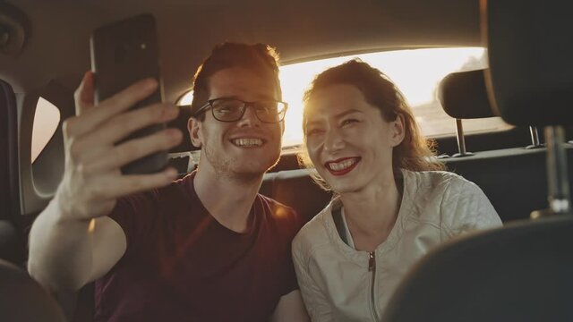 Young Male Blogger Making Selfie With Charming Asian Girl On Phone Smartphone In Back Seat Of Car At Sunset. Friends Smiling On Camera. Handsome Man In Glasses Travelling By Car. Concept Of Roadtrip