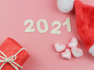 flat lay of wooden number 2021  on pink background with Christmas hat, gift boxes and heart shape marshmallow.