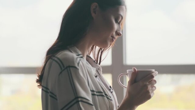 Positive pregnant woman looking at the window while drinking coffee indoors at home