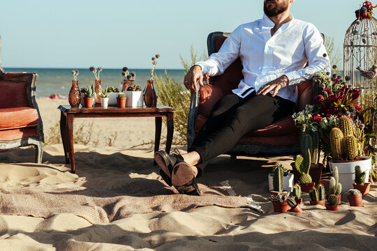 Cropped photo of the groom sitting on the chair on the wedding ceremony on the beach. relaxed man in a cozy setting for a date. wedding decoration with cactus and succulents on the seaside.