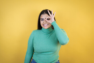 Young caucasian woman over isolated yellow background doing ok gesture shocked with smiling face, eye looking through fingers