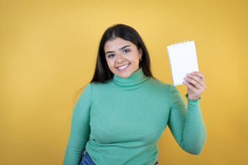 Young caucasian woman over isolated yellow background smiling and showing blank notebook in her hand