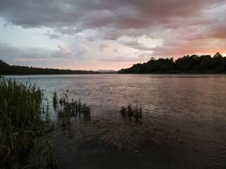 Storm clouds with rain, summer shower. Landscape with river, dark clouds, tall water grass and mountain in distance at sunset. Dramatic riverine scene during cloudy dusk.