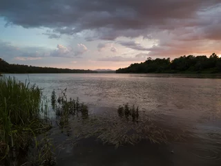 Foto op Canvas Storm clouds with rain, summer shower. Landscape with river, dark clouds, tall water grass and mountain in distance at sunset. Dramatic riverine scene during cloudy dusk. © slobodan