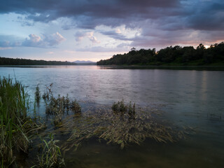 Storm clouds with rain, summer shower. Landscape with river, dark clouds, tall water grass and mountain in distance at sunset. Calm riverine scene during cloudy evening.
