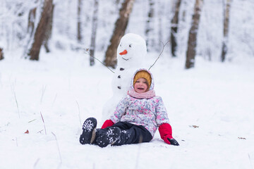 Fototapeta na wymiar Happy child plays with a snowman on a winter walk in nature in the forest. Funny little girl laughs and sits on the snow next to a snowman in winter outdoors