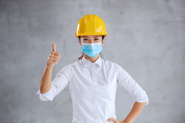 Young female architect with helmet on head, with protective mask standing indoors and holding thumbs up.