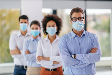 Businessman with face mask standing in corporate firm with arms crossed.Behind him are his colleagues with arms crossed and face masks, too. Although it's corona business can't stop.