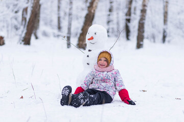 
Happy child plays with a snowman on a winter walk in nature in the forest. Funny little girl laughs and sits on the snow next to a snowman in winter outdoors