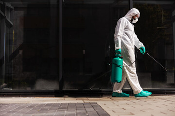 Side view of man in sterile protective uniform with rubber gloves holding sprayer with disinfectant and spraying outdoors in order to prevent corona virus form spreading.