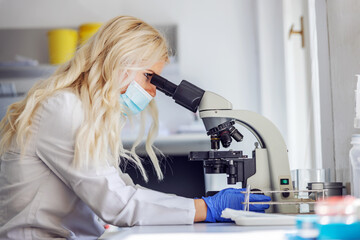 Side view of attractive blond female lab assistant with rubber gloves and face mask sitting in...