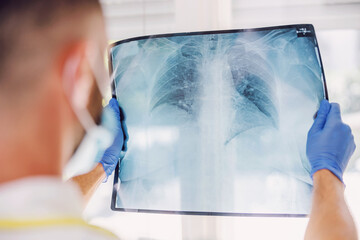 Rear view of doctor with face mask and rubber gloves standing in hospital and looking at x-ray of...