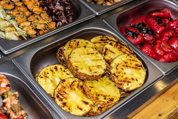 A metal bowl contains a large number of slices of pineapple cut into rings and grilled. Delicious food cooked over an open fire, which is offered at a street food fair, event, festival.