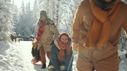Two young men running and pulling sleds with joyous girlfriends riding on them while having fun in forest on sunny winter day