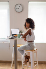Casual comfortable young black woman taking a snack and working from home with her laptop.