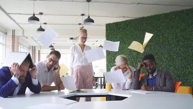 Angry unbalanced female boss scream, scolds employees for not fulfilling the plan at work. Frightened workers sit quietly, listening to her swearing, she scatters documents.