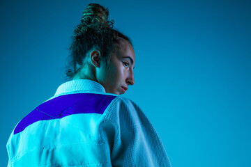 Fototapeta premium Posing. Professional female judoist in white judo kimono practicing and training isolated on blue neoned studio background. Grace of motion and action. Healthy lifestyle, sport and movement concept