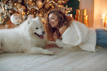 A beautiful woman with a white Samoyed, hugs, against the background of a Christmas tree with candles in a decorated room in Christmas. Happy New Year. High quality photo.