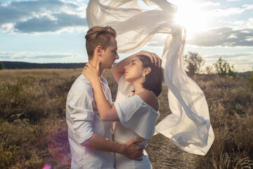 portrait of a young wedding couple, a guy and a girl of twenty-two years old, in a field with a flying white ribbon behind, against the backdrop of the setting sun, cloudy blue sky. - Powered by Adobe