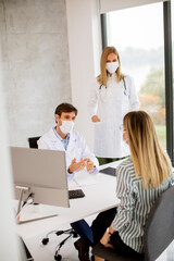 Medical couple doctors with protective medical masks talking with female patient and using computer in the office