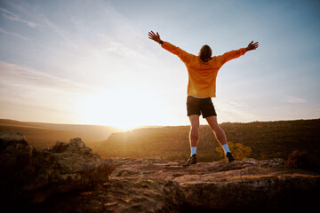 Rear view of young man standing with arms outstretched at mountain top during morning sunrise. conquering adversity through motivation and determination