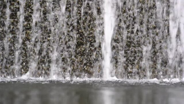 Water falling pouring into a poolside from stone wall, close up. Background with running water. Waterfall fountain stri and spray drops stone surface.