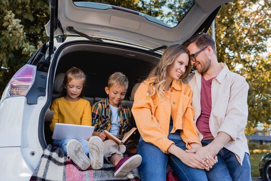 Smiling kids with book and digital tablet sitting in truck of car near parents holding hands outdoors