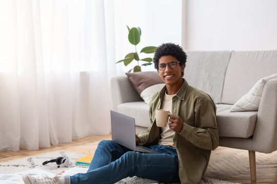 Positive Black Teen Guy Sitting On Floor With Laptop And Cup Of Coffee, Studying Online From Home, Copy Space