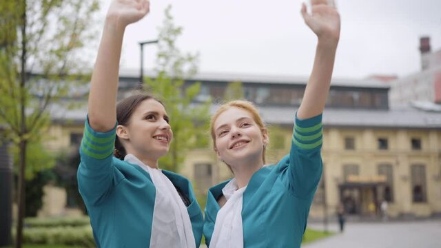 Positive young slim women in stewardess uniform waving and looking up. Portrait of smiling elegant Caucasian air hostesses standing outdoors. Travelling and tourism concept.
