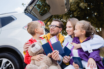 Smiling parents hugging kids with passports and air tickets near car outdoors
