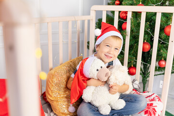 a child in a cap is sitting in a crib in a children's room with a new year's interior near the Christmas tree and playing with soft bears