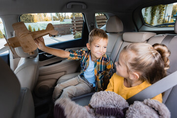 Boy with toy plane looking at sister with soft toy on blurred foreground in car