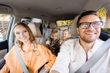 Cheerful kids pointing with fingers near parents on blurred foreground in car