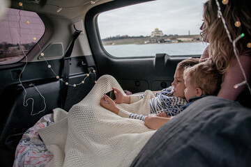 Family, mother and son in warm clothes relaxing in the car parked in the nature. People dressed sweater and hats   take a rest in automobile,covered with a blanket in garland decorated car