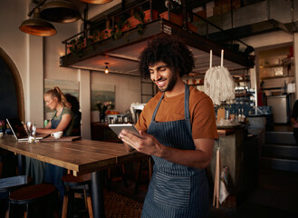 Male waiter with curly hair working in cafe wearing apron with using digital tablet with female...