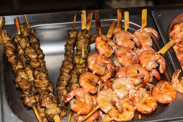 A large amount of freshly prepared seafood, skewered on wooden skewers, is in a metal bowl. Delicious food cooked over an open fire, which is offered at a street food fair, event, festival.