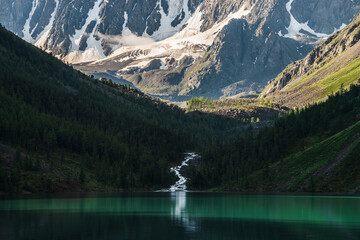 Beautiful big glacier in sunlight, rocky snowy mountains, low cloud, coniferous forest on hills, mountain lake and highland creek reflected in clear water. Atmospheric alpine scenery at sunny morning.