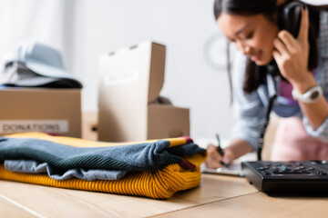 Clothes on table near asian woman talking on telephone near boxes on blurred background in charity center