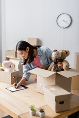 Fototapeta premium Smiling asian volunteer writing on clipboard near packages with soft toys in charity center