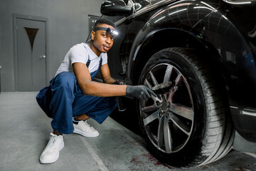 Professional African male worker in overalls and black protective gloves, cleaning alloy wheels rims of luxury car with a special brush for cast wheels in a detailing workshop, looking at camera