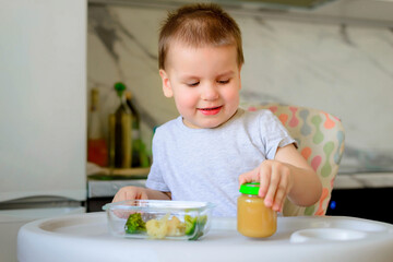 Happy 2-year-old boy kid eats cauliflower, broccoli and mashed potatoes in a jar in his kitchen at home. Baby food, solid food concept.