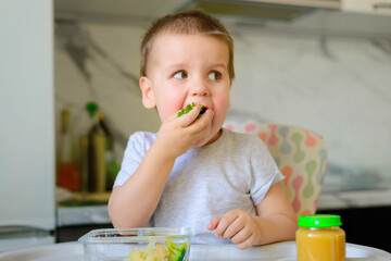 Happy 2-year-old boy kid eats cauliflower, broccoli and mashed potatoes in a jar in his kitchen at home. Baby food, solid food concept.