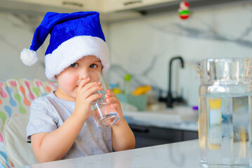 Funny boy kid in a blue Santa hat drinking filtered water from a glass in the kitchen. Holidays, health concept.