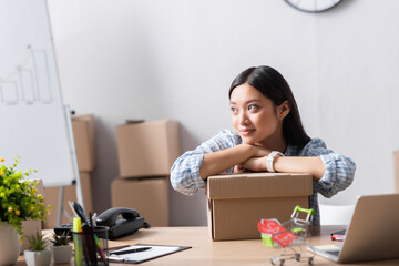 Smiling asian volunteer looking away while leaning on carton box on desk in charity center on blurred foreground