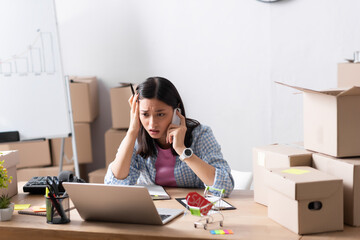 Worried asian volunteer talking on smartphone while looking at laptop at desk with stationery and carton boxes in charity center