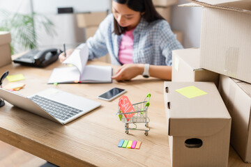 Price tag with sale lettering in shopping cart near carton boxes on desk with blurred asian volunteer on background