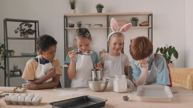 Diverse school kids wearing aprons standing at table in kitchen, smiling, talking, cooking dough. Mixed-Race girl pouring flour into bowl trough sifter that child with bunny-ears headband holding